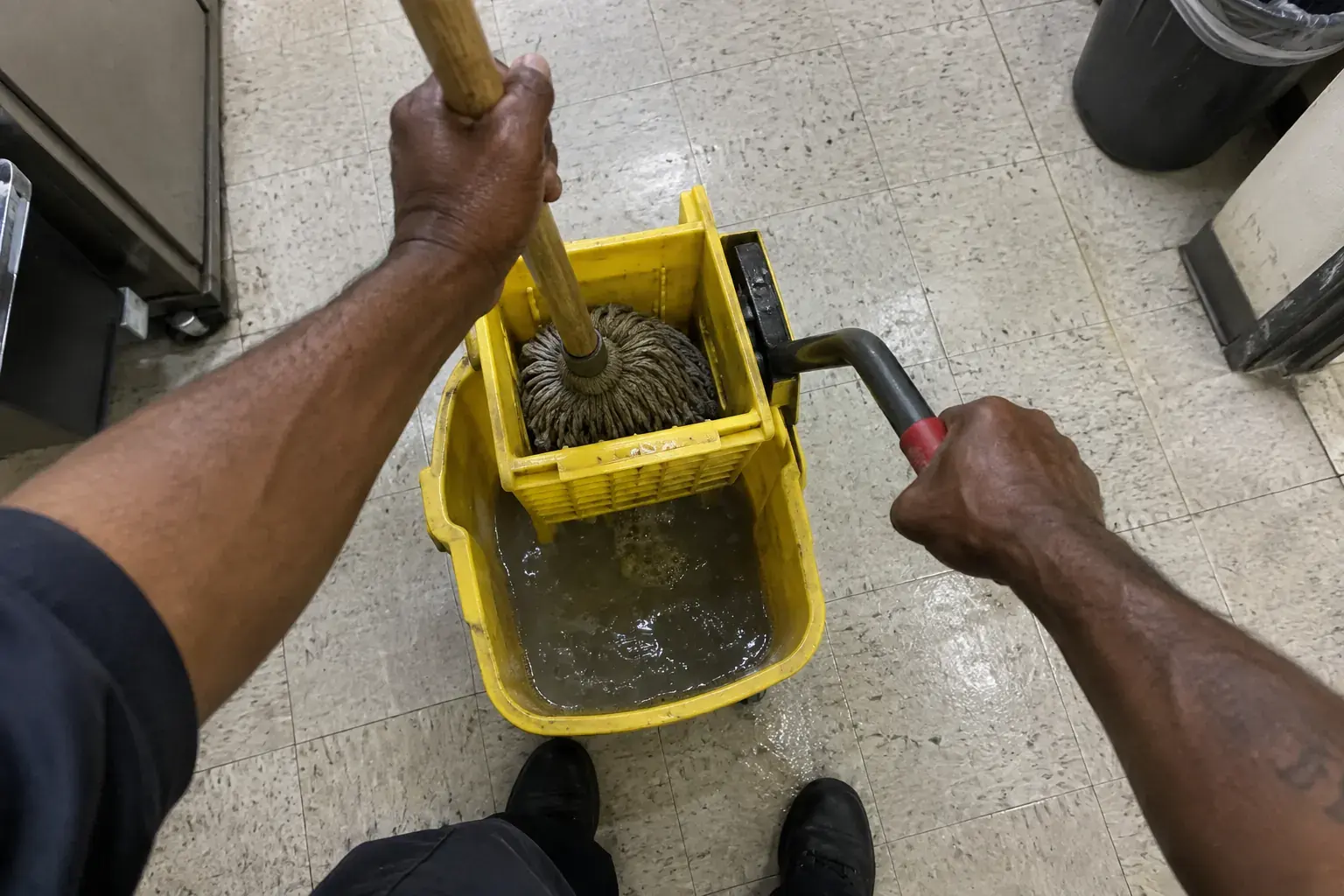 First-person view of a cleaner wringing a string mop in a yellow bucket, both hands in view.