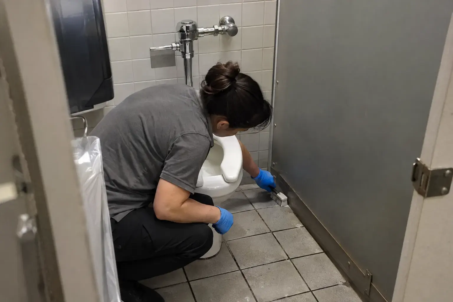 A cleaner in gloves crouched in a tight restroom stall, scrubbing the floor corner behind a toilet with a small brush.