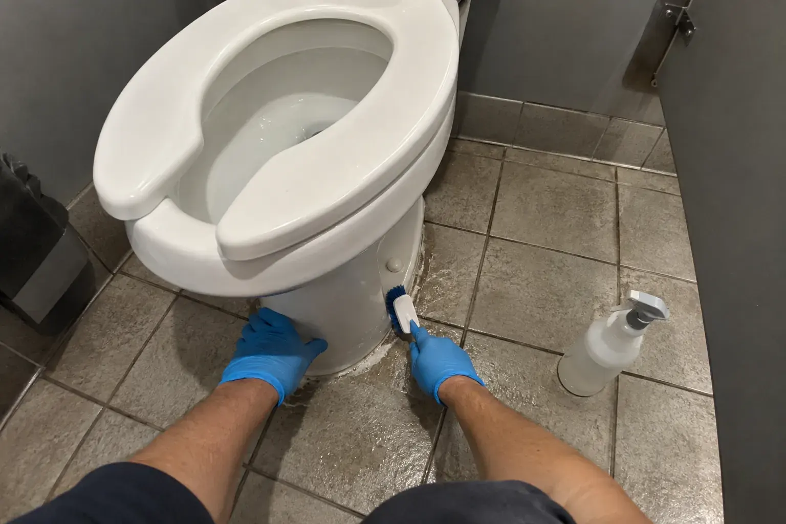First-person view of gloved hands scrubbing around the base of a toilet in a tight stall.