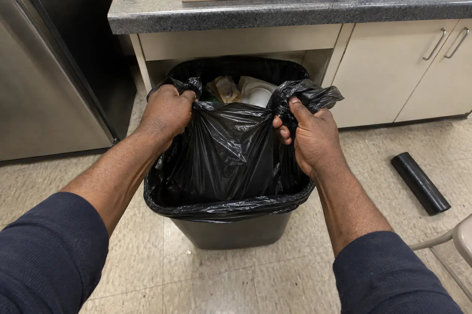 First-person view of a cleaner pulling a full trash liner out of a break-room can with both hands.